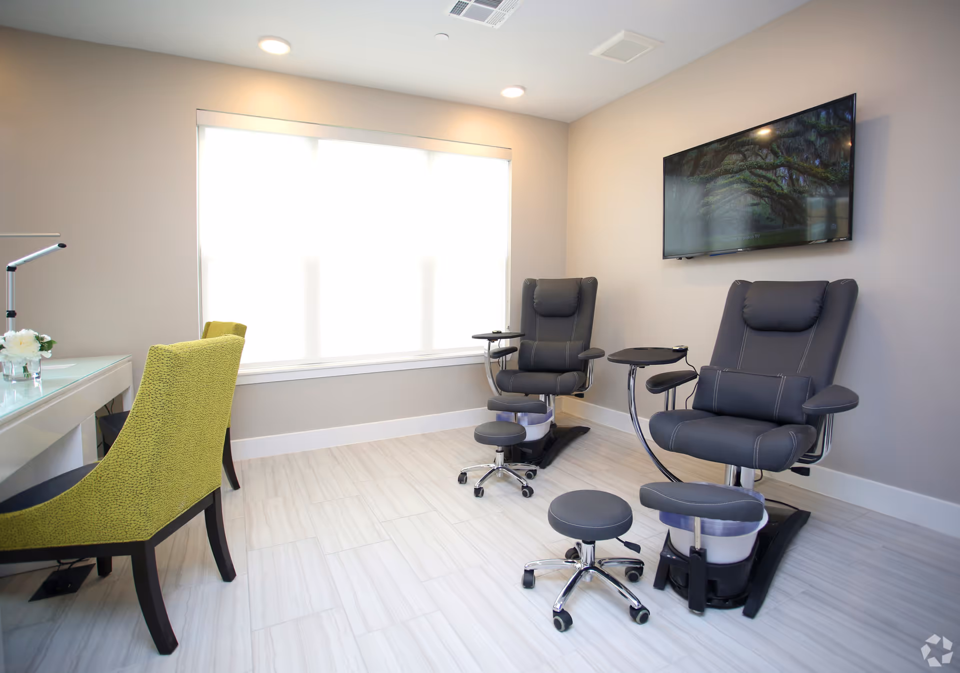 A bright room with two black pedicure chairs equipped with footrests and small attached tables, a large window with blinds, a wall-mounted flat-screen TV, and a green upholstered chair next to a white desk with a lamp and small flower arrangement.