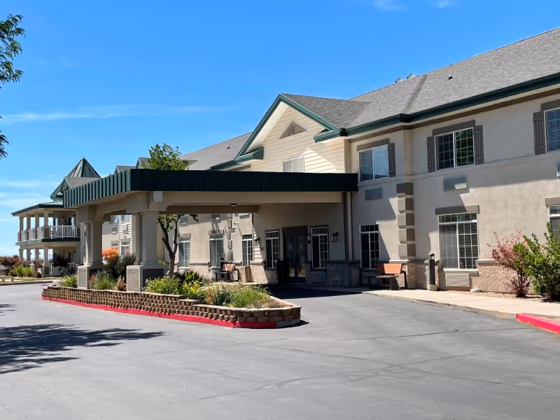 Front entrance with a covered porte-cochère of a two-story beige assisted living building, landscaped beds and a paved driveway.