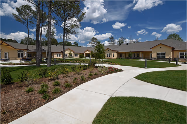 Single-story yellow senior living building surrounding a landscaped courtyard with paved walkways, pine trees, and a blue sky.