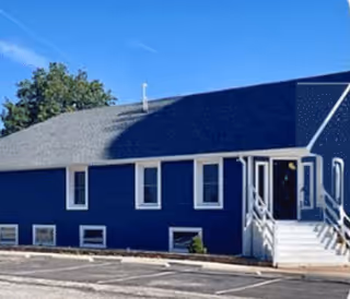 Exterior view of a single-story blue building with white trim, several windows, and a white staircase leading to a door. The building is situated next to a paved parking area with some greenery and trees in the background.