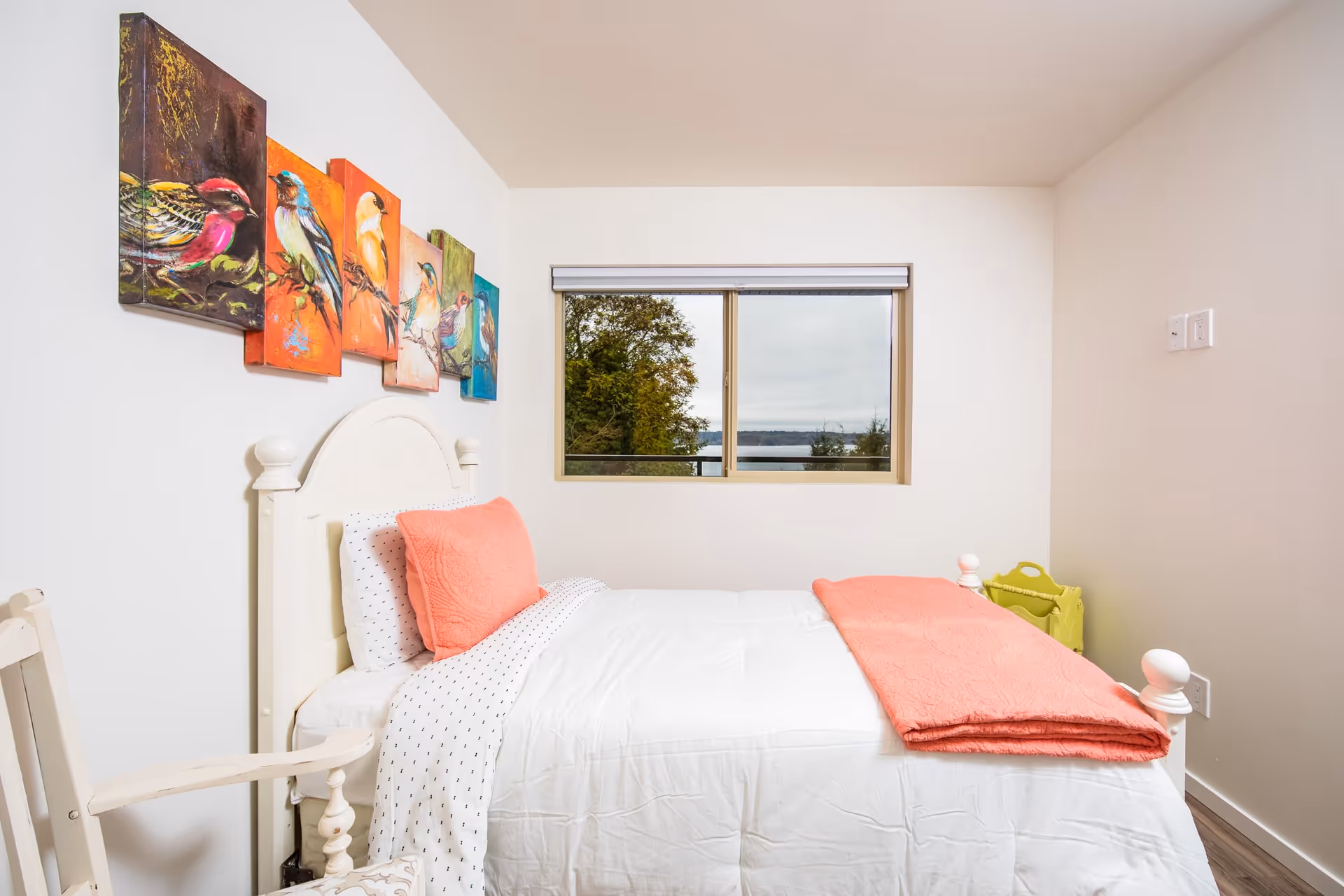 A bright bedroom with a white bed featuring coral-colored pillows and a matching blanket. Above the bed is a colorful wall art of various birds. A window shows a view of trees and a body of water outside. A white chair is partially visible on the left side of the image.