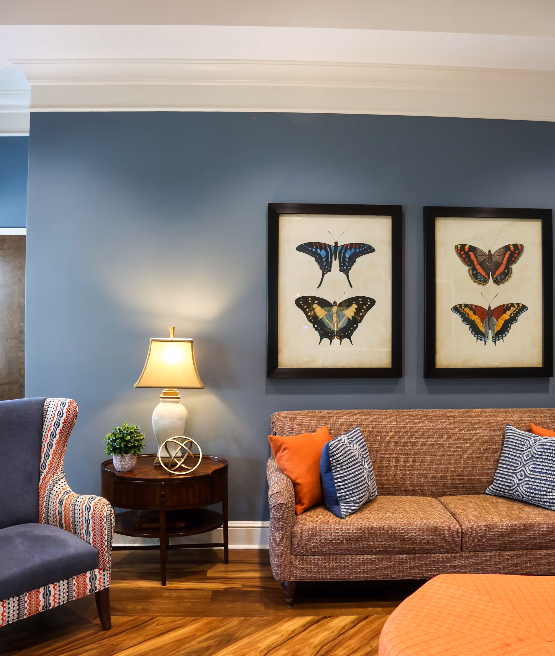 A cozy living room area with a brown textured sofa adorned with orange and patterned blue pillows. Next to the sofa is a round wooden side table with a white lamp, a small green plant, and a decorative gold orb. On the left side, there is a patterned armchair with blue, orange, and white colors. The wall behind is painted blue and features two framed butterfly artworks.