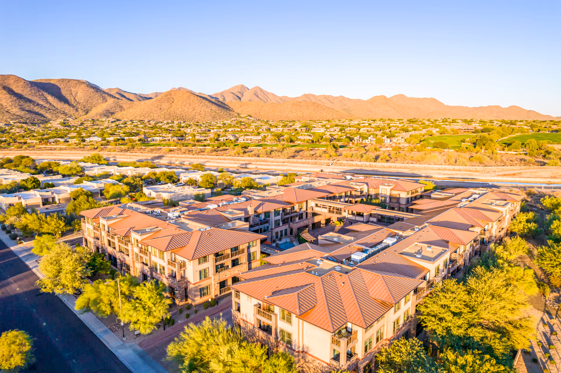 Aerial view of Via Linda Assisted Living facility with multiple connected buildings featuring terracotta roofs, surrounded by trees and roads, with desert mountains and a clear blue sky in the background.