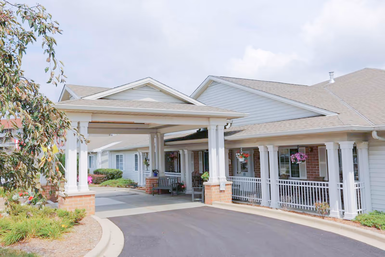 Exterior view of Brenwood Park Assisted Living facility showing a covered entrance with white columns and a paved driveway. The building has light-colored siding, a sloped roof, hanging flower baskets, and some greenery around the entrance.