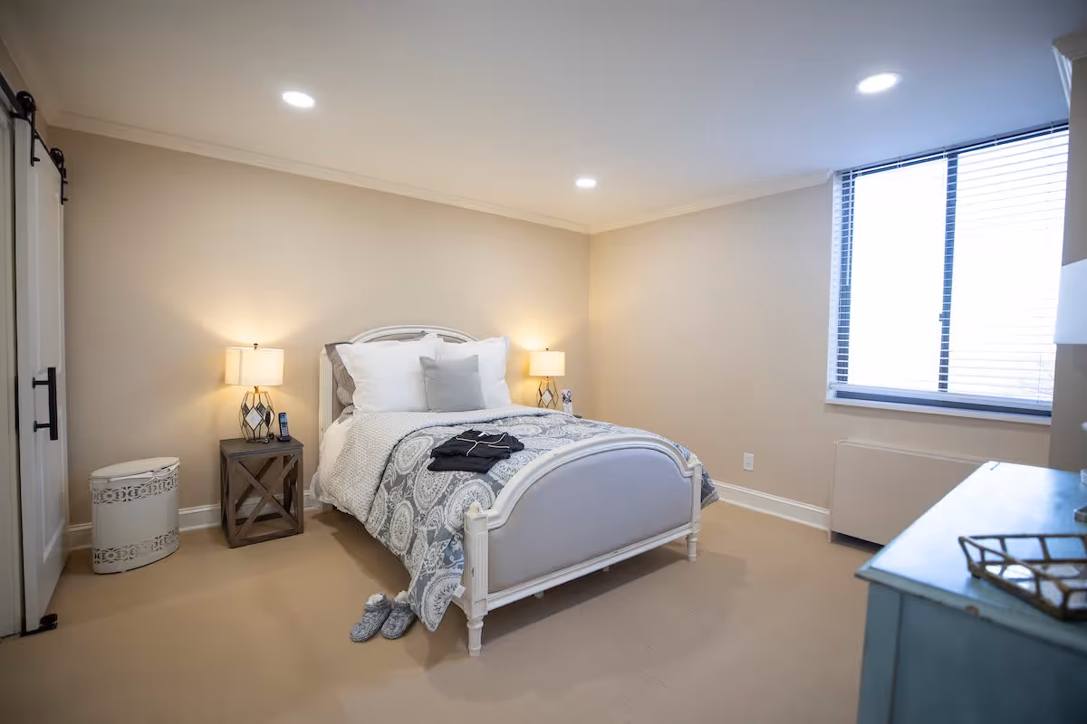 A cozy bedroom with a white bed frame and a patterned gray and white comforter. There are two bedside tables with lamps on each side of the bed. A pair of slippers is placed on the floor near the foot of the bed. The room has beige walls, a window with blinds, and a blue dresser on the right side.