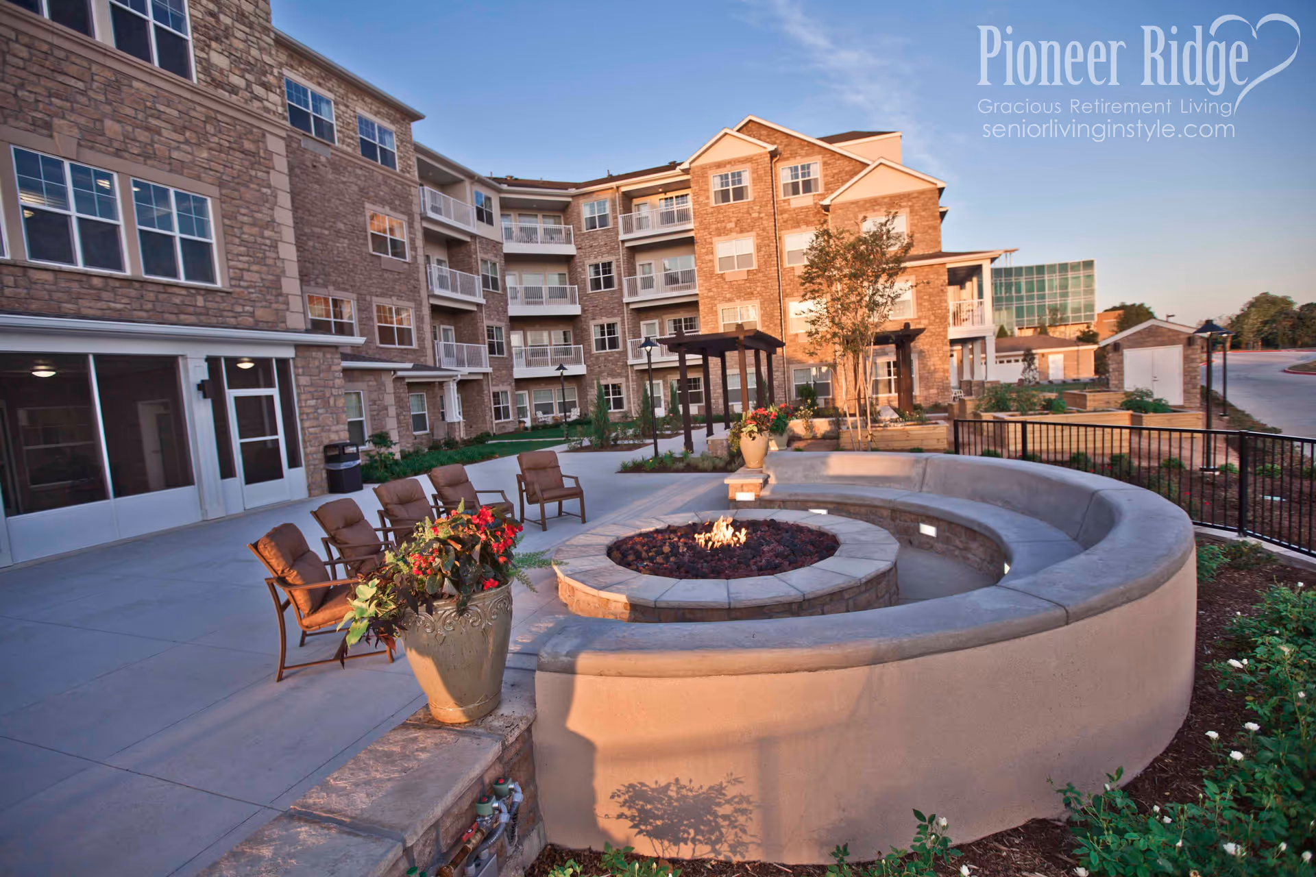 Outdoor patio area of a senior living facility with a circular stone fire pit surrounded by a curved concrete bench and several cushioned chairs. The background shows a multi-story building with stone and brick exterior, balconies, and windows. There are potted plants with flowers and landscaped greenery around the patio. The sky is clear and the scene is lit by natural sunlight.