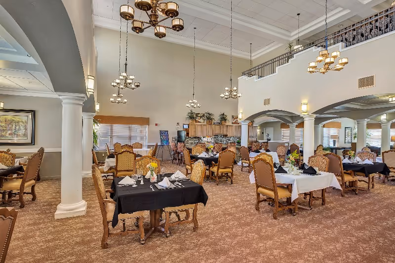 Spacious dining room in Sierra Del Sol Assisted Living with multiple tables set for meals, featuring elegant chairs, black and white tablecloths, floral centerpieces, and hanging chandeliers. The room has high ceilings, large windows with blinds, decorative columns, and warm lighting.