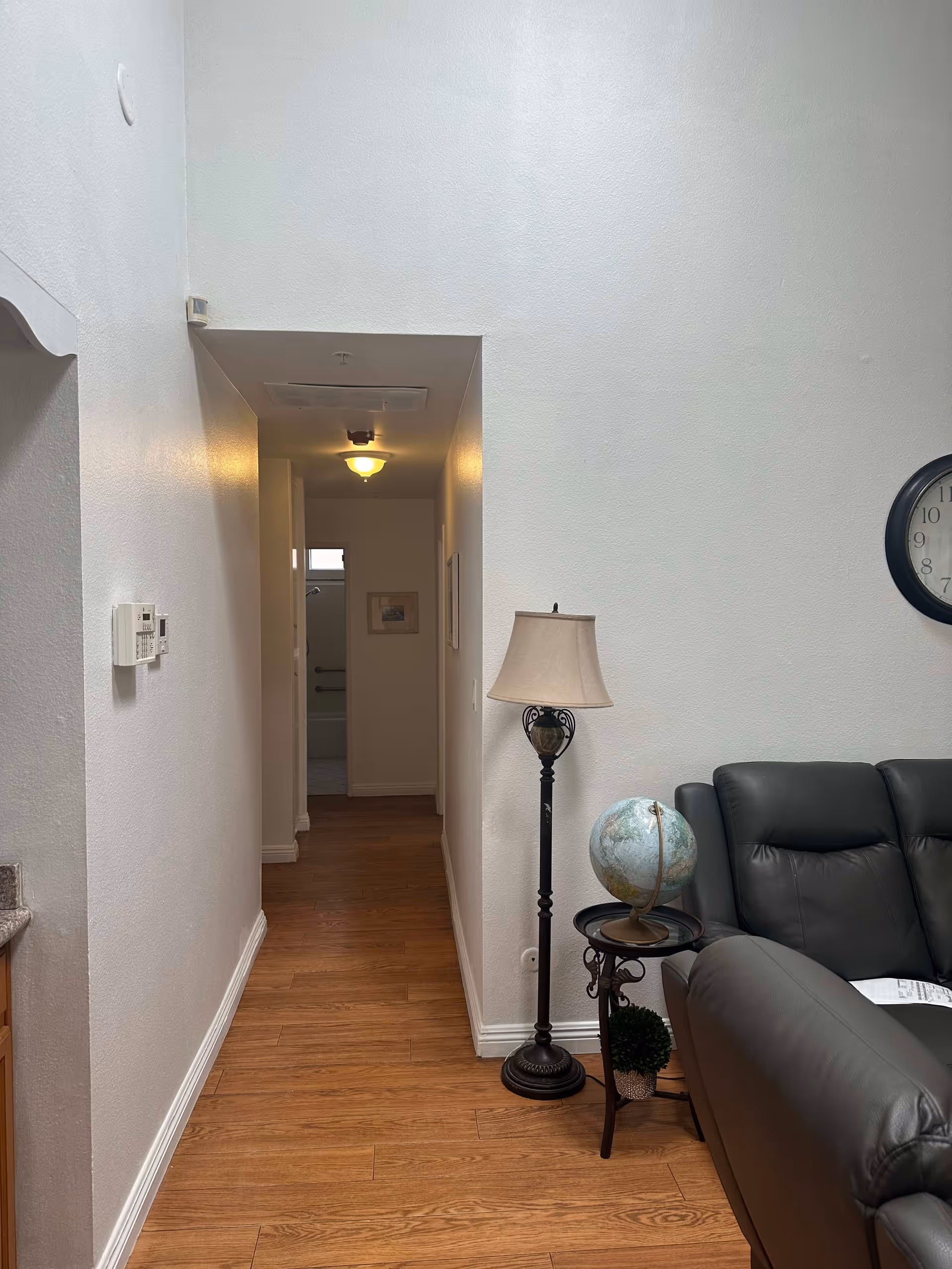Interior view of a hallway in a home care facility with wooden flooring and white walls. On the right side, there is a black leather couch, a small round table with a globe and a small plant, and a tall floor lamp with a beige lampshade. A round wall clock is partially visible above the couch. The hallway leads to a bathroom with a visible bathtub and grab bars.