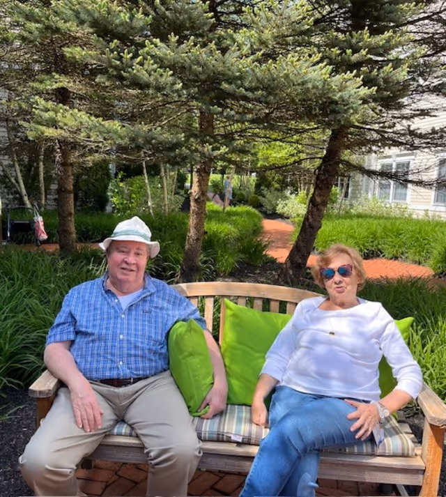 An elderly man and woman sitting on a wooden bench with green cushions in a garden area with trees and shrubs. The man is wearing a white hat, blue checkered shirt, and beige pants. The woman is wearing sunglasses, a white long-sleeve shirt, and blue jeans. They are outdoors on a sunny day with a brick pathway and building visible in the background.