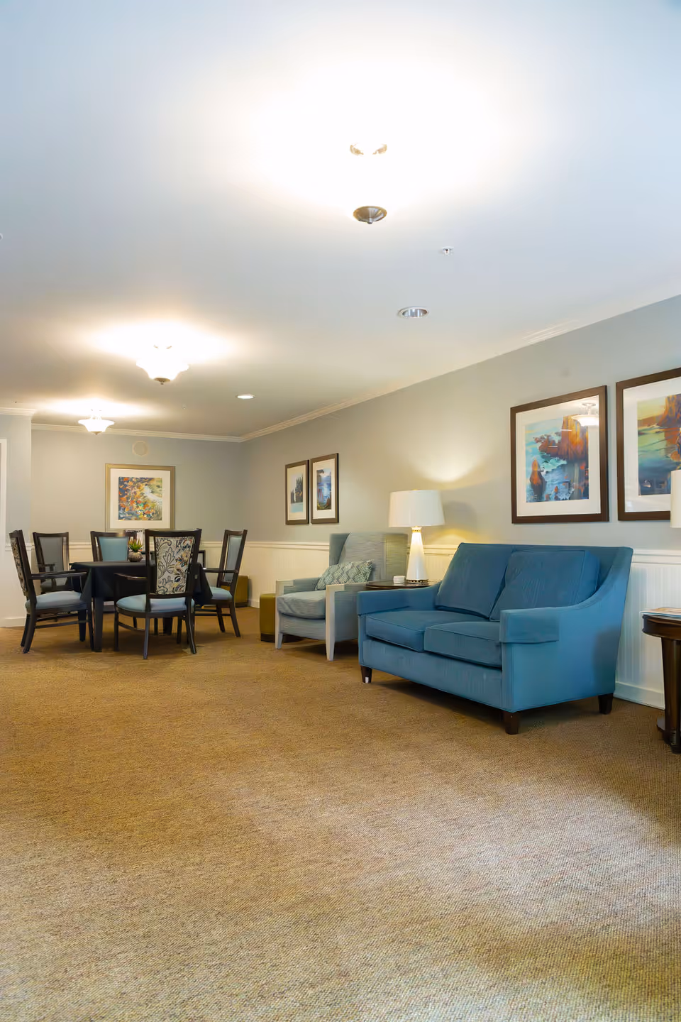 A cozy living room area with a blue loveseat, a light blue armchair, a side table with a lamp, and framed artwork on the wall. In the background, there is a dining area with a round table and six chairs, along with additional framed artwork on the walls. The room has beige carpeting and soft overhead lighting.