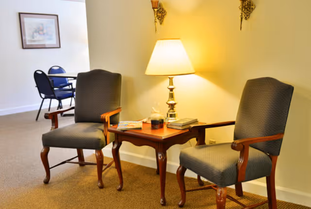 Two upholstered armchairs with wooden arms and legs are positioned on either side of a wooden side table with a lamp, books, and a decorative item. In the background, there is a small table with two chairs and a framed picture on the wall.
