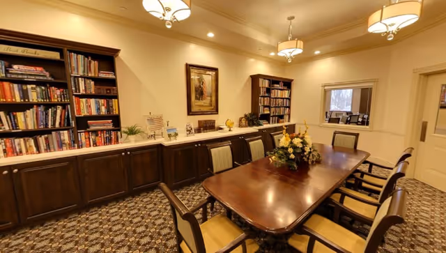 A well-lit meeting or dining room with a long wooden table surrounded by eight chairs. The room features built-in dark wood bookshelves filled with books and decorative items along one wall. There are three ceiling light fixtures and a floral centerpiece on the table. A window and a door are visible on the right side of the room.