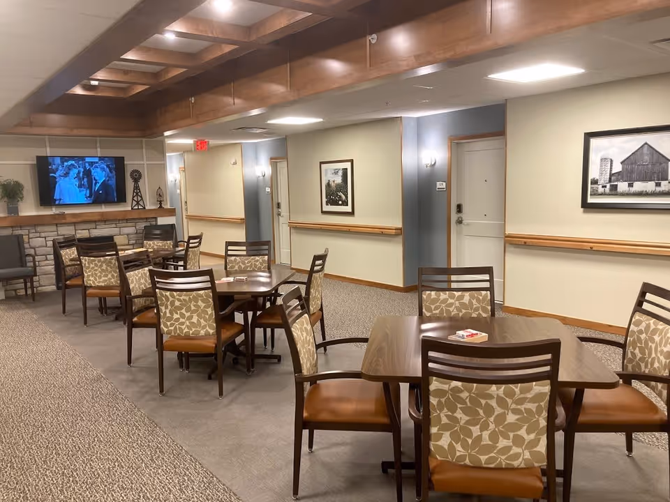 Interior view of a senior living facility common dining area with several wooden tables and chairs featuring patterned upholstery. A flat-screen TV is mounted on the far wall above a stone fireplace. The walls are decorated with framed pictures, and the ceiling has wooden beams with recessed lighting.