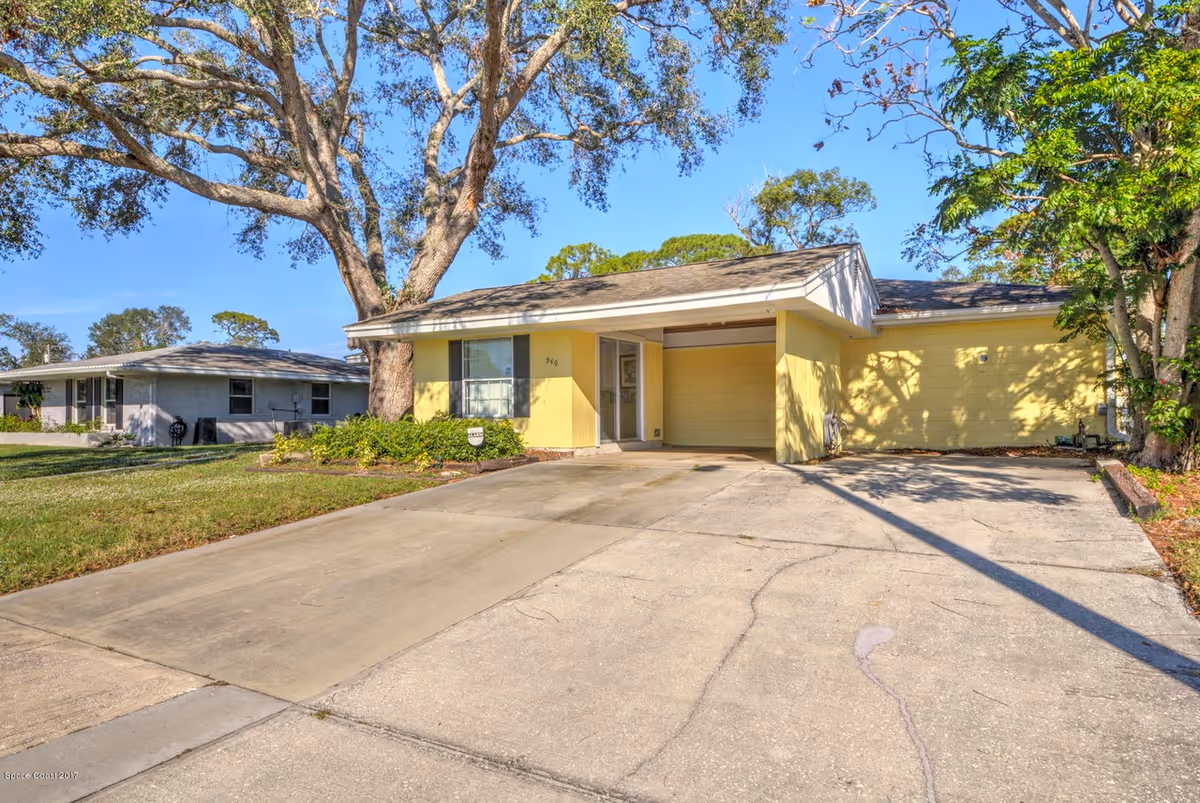 A yellow, single-story building with a carport, driveway, and large trees in front.