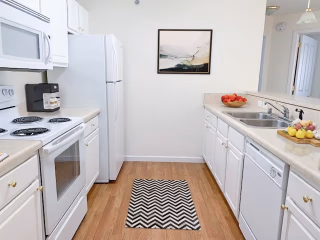 Bright galley kitchen with white cabinets and appliances, a double sink, countertop fruit bowls, and a chevron rug on hardwood floor.