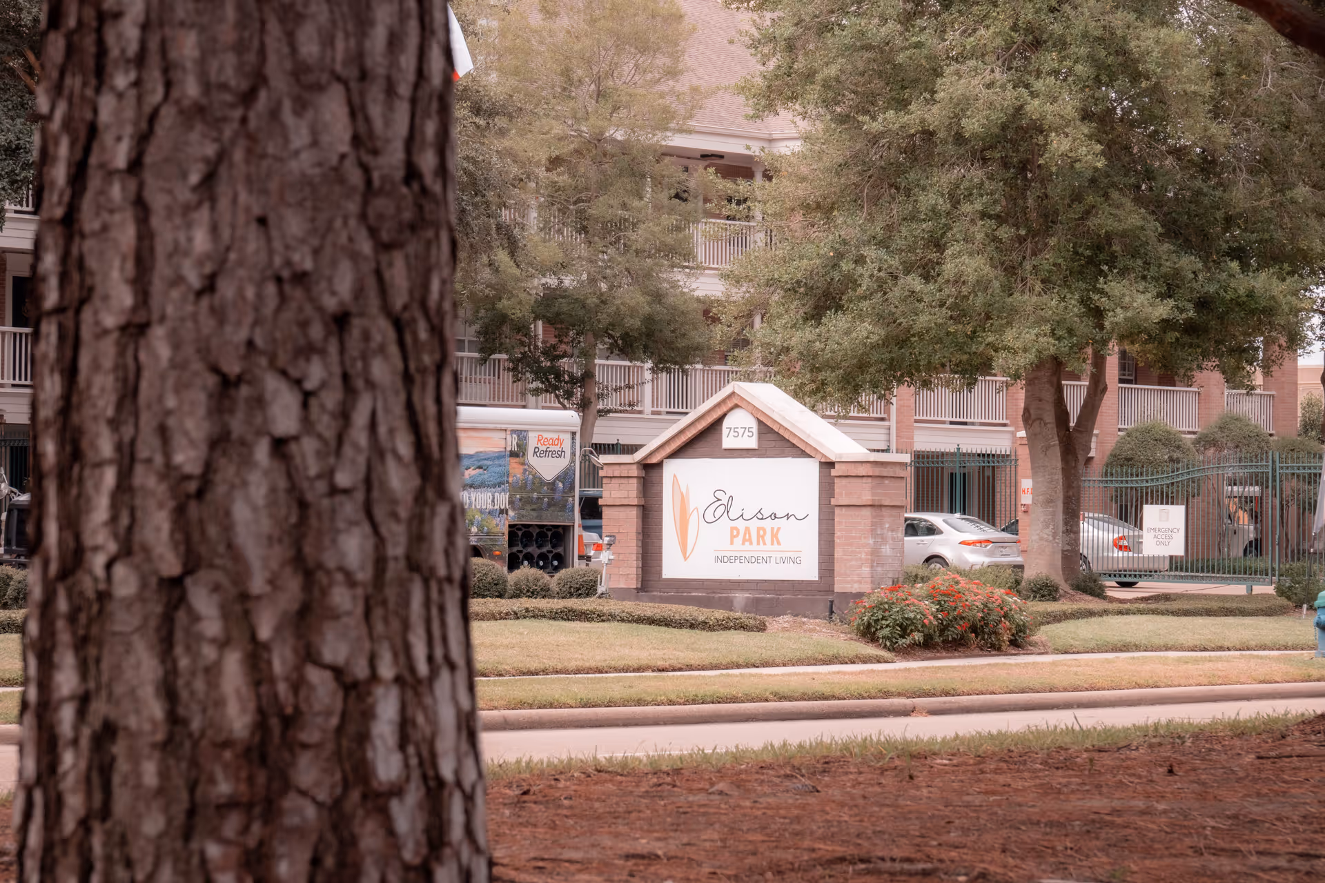View of the entrance sign for Elison Park Independent Living, a senior living community, with a large tree trunk in the foreground and a multi-story building with balconies and parked cars in the background.