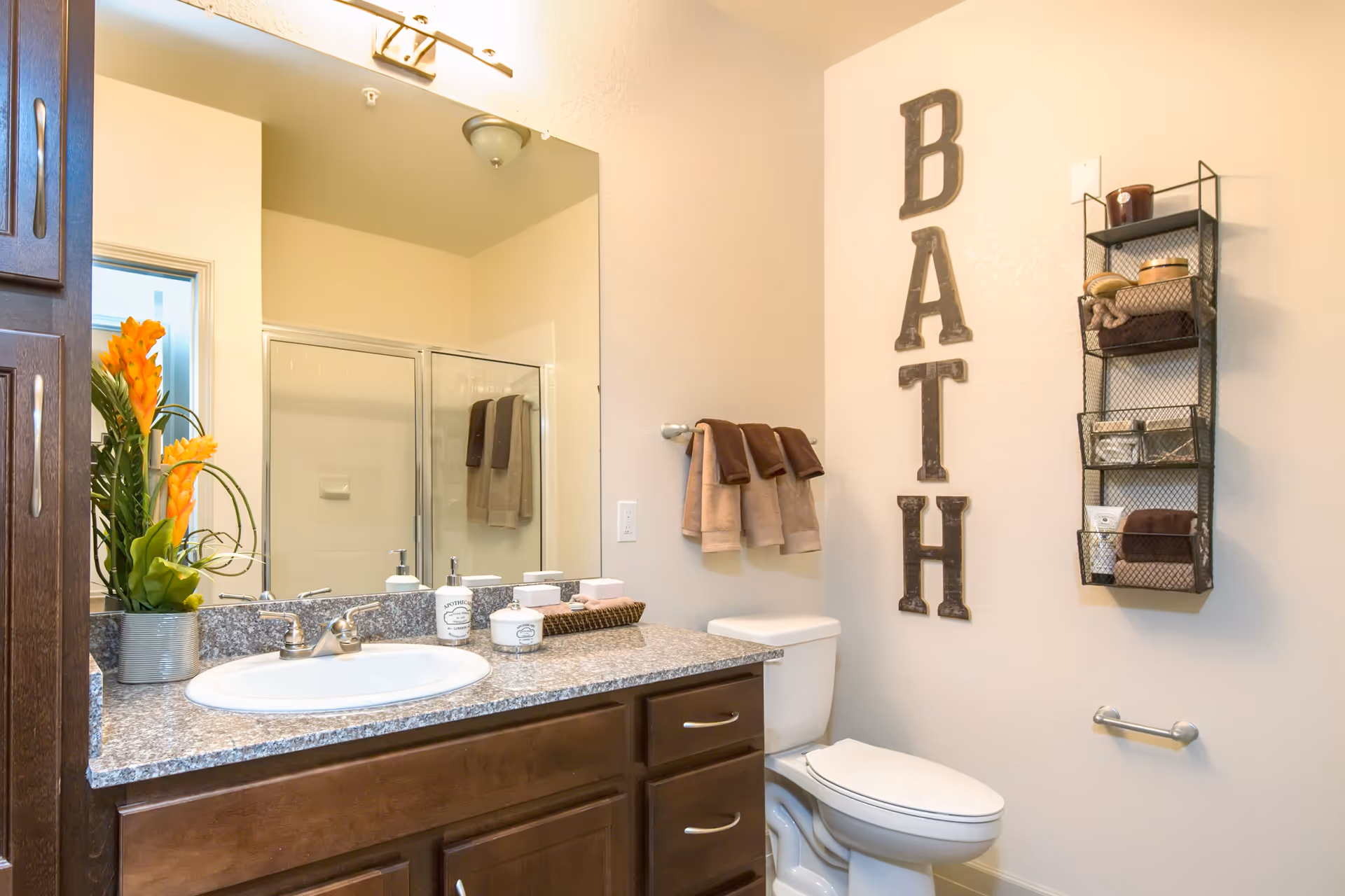 Bathroom with a granite-top vanity and sink, large mirror, toilet, glass shower, wall-mounted shelf, and decorative 'BATH' letters on the wall.
