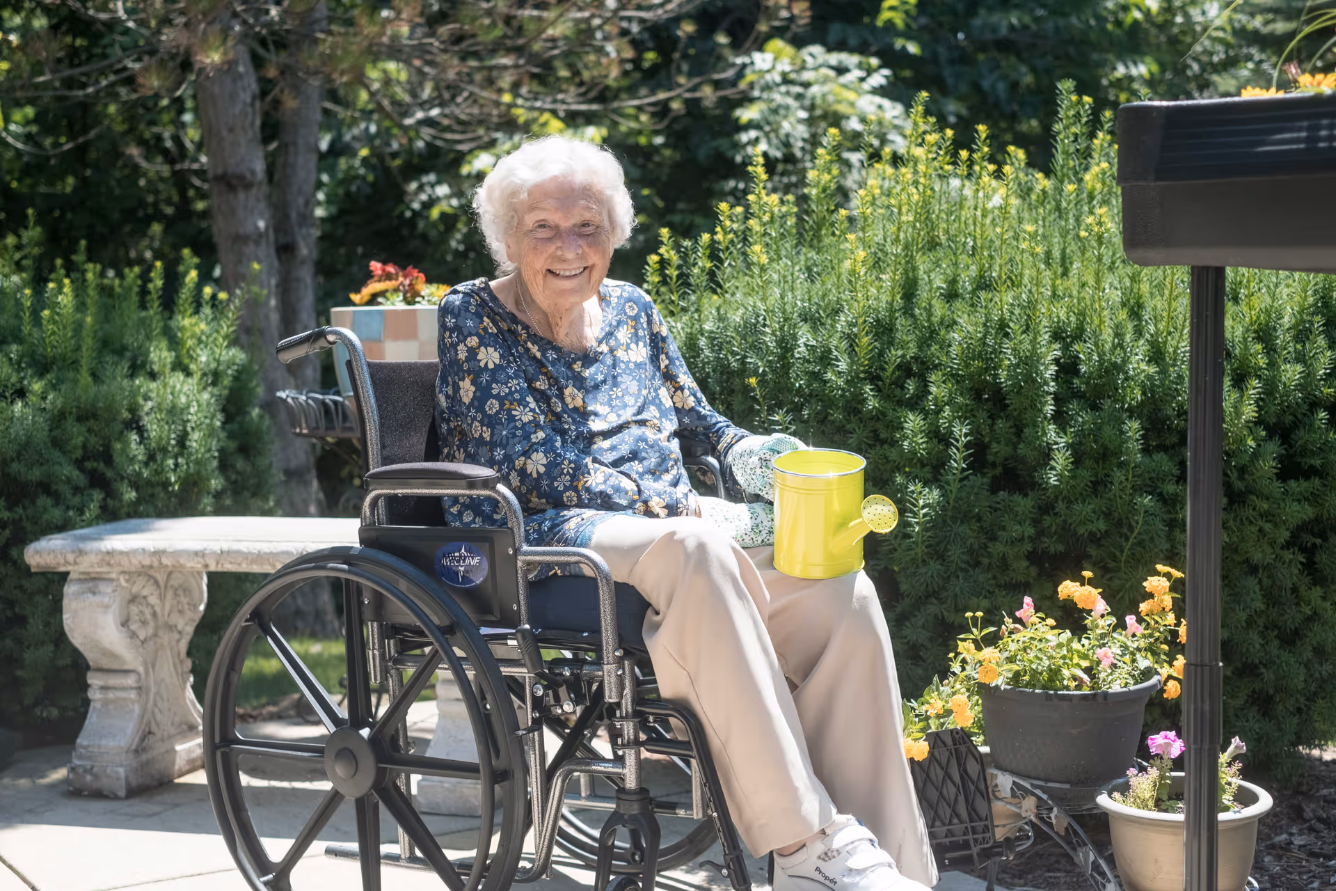 An elderly woman sitting in a wheelchair outdoors, smiling and holding a yellow watering can. She is surrounded by green bushes and potted flowers, with a stone bench visible in the background.
