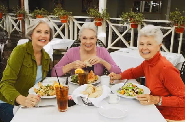 Three senior women smiling while seated at an outdoor dining table with salads, iced tea, and a basket of bread.