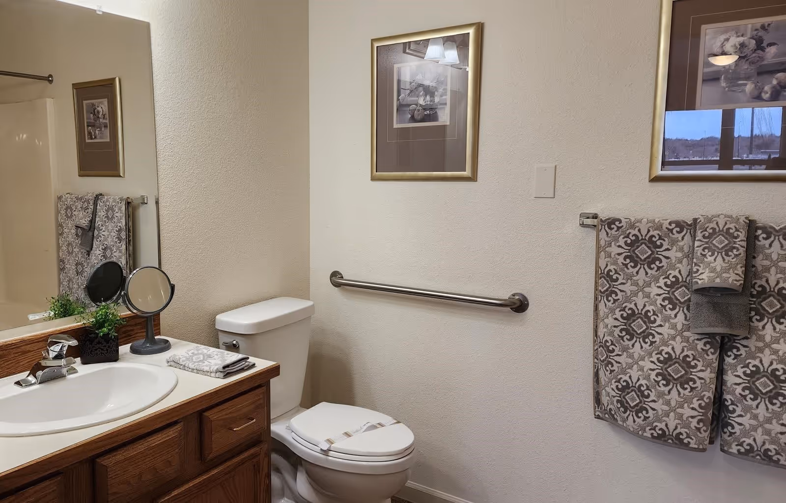 A bathroom with a white toilet and a wooden vanity with a white sink. Above the sink is a large mirror reflecting part of the shower area. On the vanity are a small plant, a round makeup mirror, and folded towels. On the wall above the toilet is a framed picture, and next to the toilet is a metal grab bar. On the right wall, there are patterned towels hanging on a towel rack and another framed picture above them.