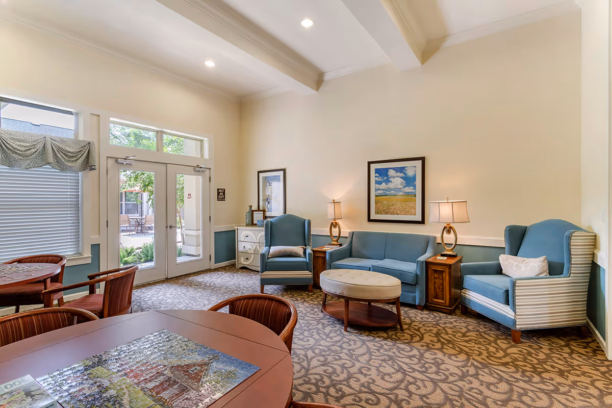 A bright and cozy living room area in an assisted living facility featuring two blue armchairs, a blue sofa, two wooden side tables with lamps, a round ottoman, and framed artwork on the wall. There are large windows and glass doors letting in natural light, with a table and chairs in the foreground.