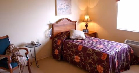 A small bedroom with a single bed covered in a floral patterned bedspread, a wooden headboard, a pillow, and a bedside table with a lamp and a small clock. There is a chair with a blanket draped over it and a small round table with a telephone. A window allows natural light into the room.