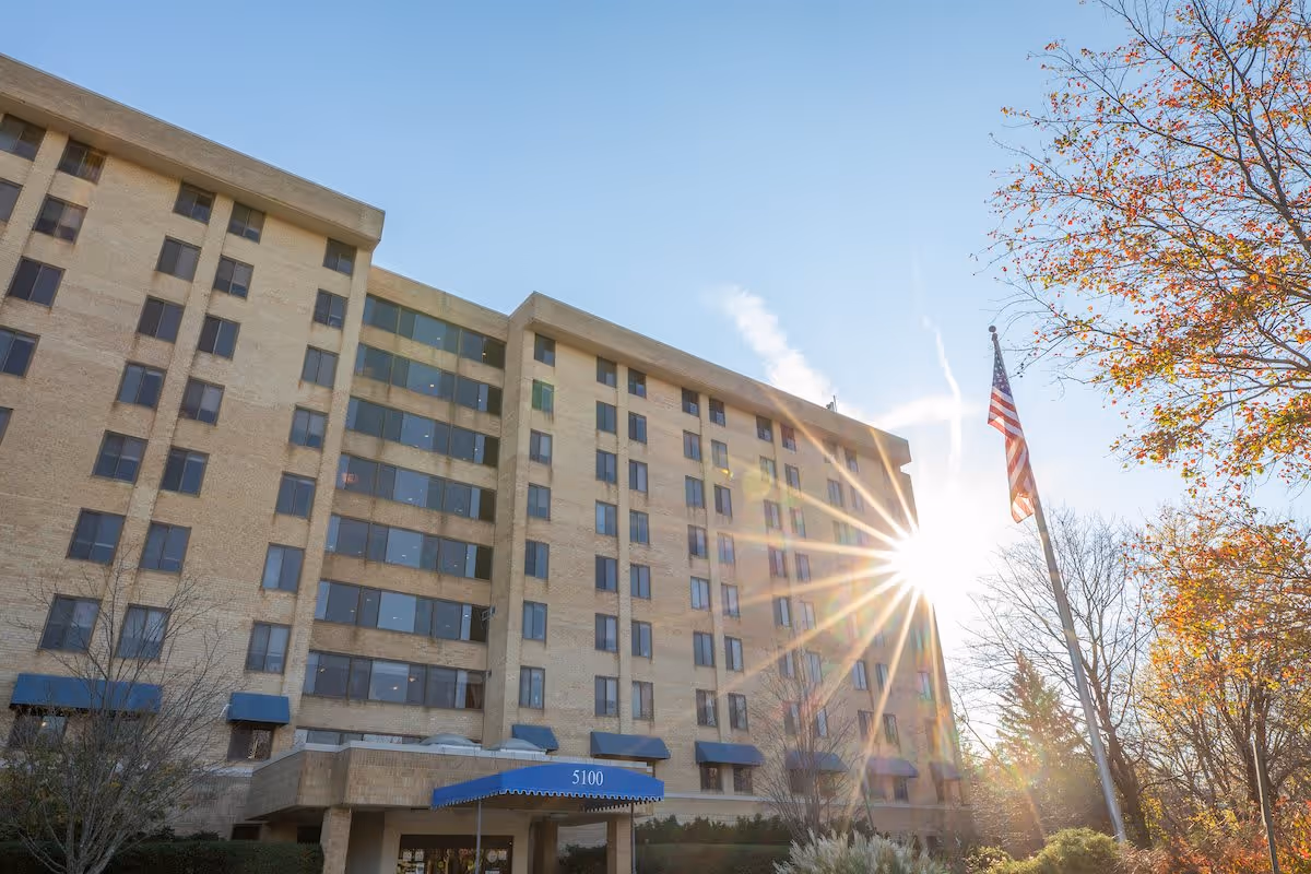 Exterior view of a multi-story senior living facility building with beige brick walls and many windows. The sun is shining brightly near the top right corner, creating a lens flare effect. An American flag is flying on a flagpole in front of the building, and there are trees with autumn foliage around the entrance area.