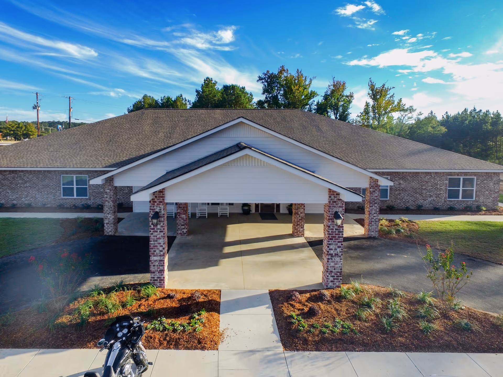 Front exterior view of a single-story brick building with a covered entrance supported by brick columns. The building is surrounded by landscaped areas with small plants and trees under a partly cloudy blue sky.