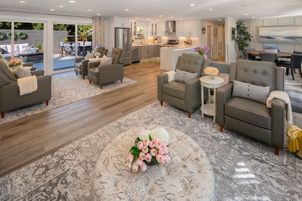 A bright and spacious senior living common area featuring comfortable gray armchairs with pillows and throws, a round ottoman with a floral arrangement, a modern open kitchen with white cabinetry and stainless steel appliances, and a dining area with a round table and chairs. Large sliding glass doors open to an outdoor patio with seating and greenery.