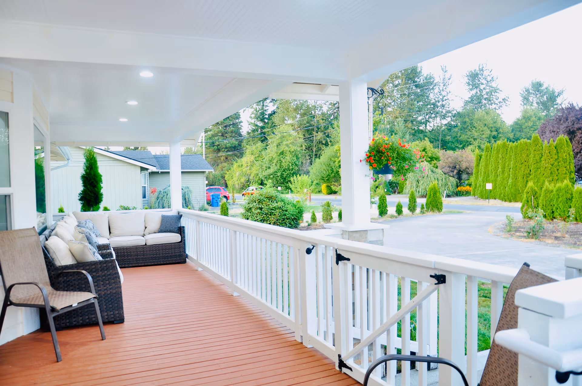 A spacious covered porch with wooden flooring and white railings. The porch is furnished with cushioned wicker sofas and chairs. There are hanging flower pots with red flowers and a view of a driveway and green trees in the background.