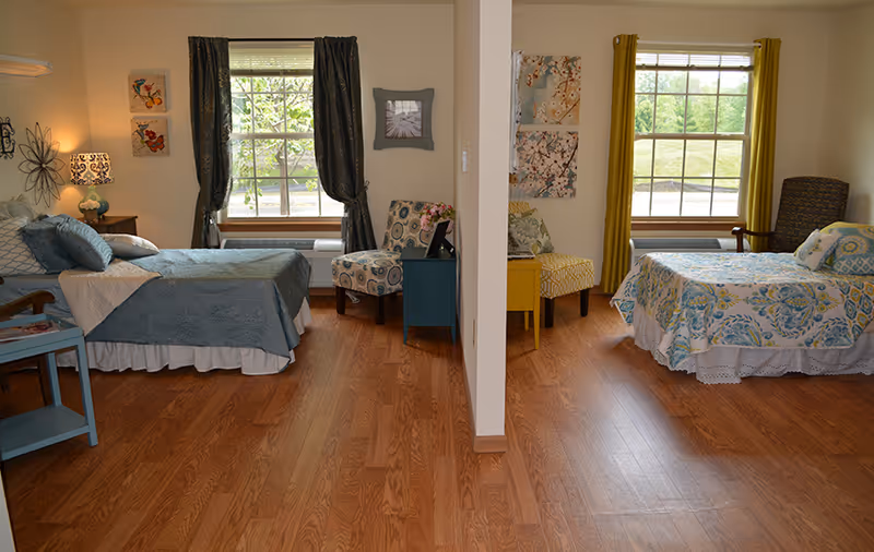 Two adjacent furnished bedrooms in an assisted living suite with hardwood floors, windows, and colorful bedding.