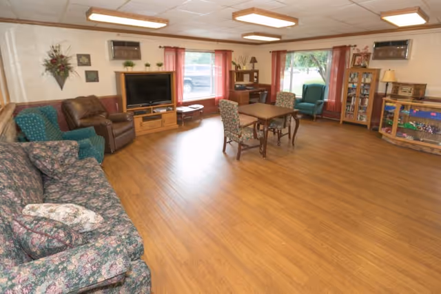 Spacious senior living common room with sofas, armchairs, a TV, and a table on wood floors.