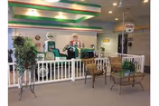 Indoor area resembling a small-scale vintage gas station display with old-fashioned gas pumps behind a white picket fence. In front of the fence, there are two wooden chairs and a glass-top table with a potted plant. The ceiling has green and white striped accents, and there are additional plants and vintage signs in the background.