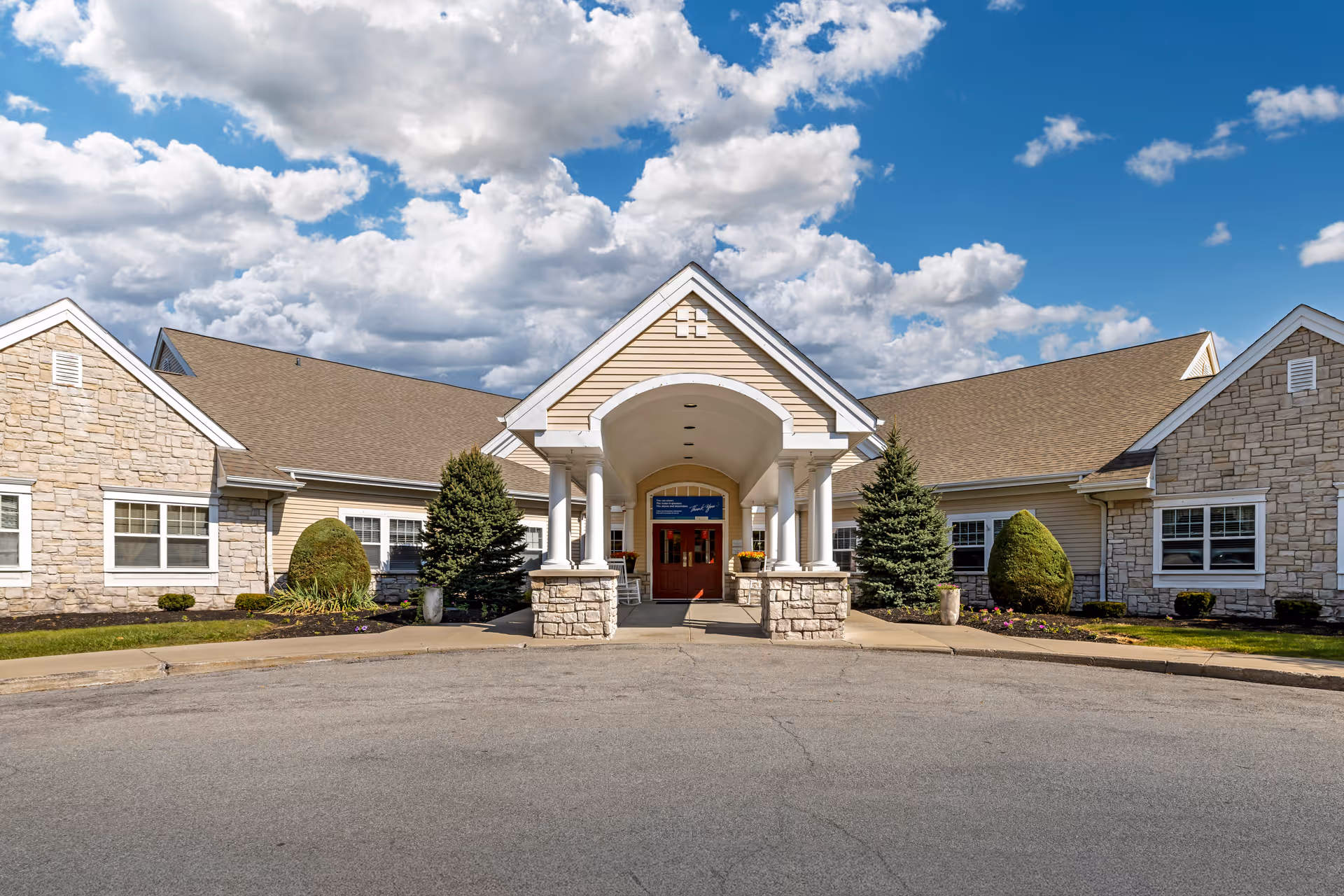 Front exterior view of Brookdale Williamsville facility with a covered entrance supported by white columns, stone and beige siding walls, manicured shrubs, and a partly cloudy blue sky.
