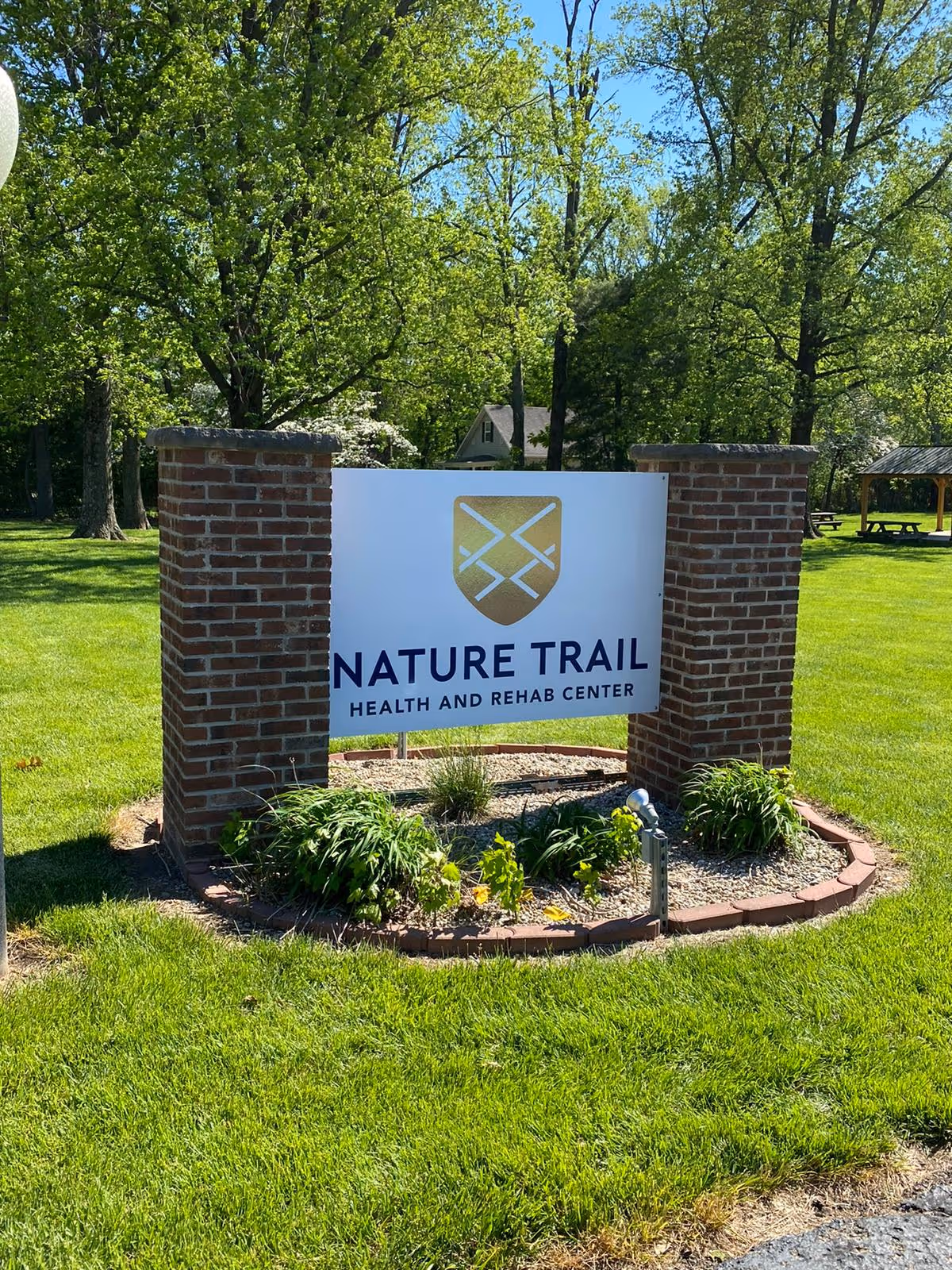 A sign for Nature Trail Health and Rehab Center mounted between two brick pillars, surrounded by green grass, plants, and trees under a clear blue sky.
