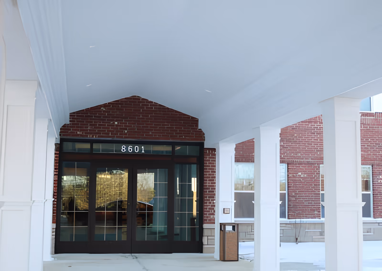 Entrance of a building with white pillars and a covered walkway leading to glass double doors with the number 8601 above them. The building exterior is made of red brick.