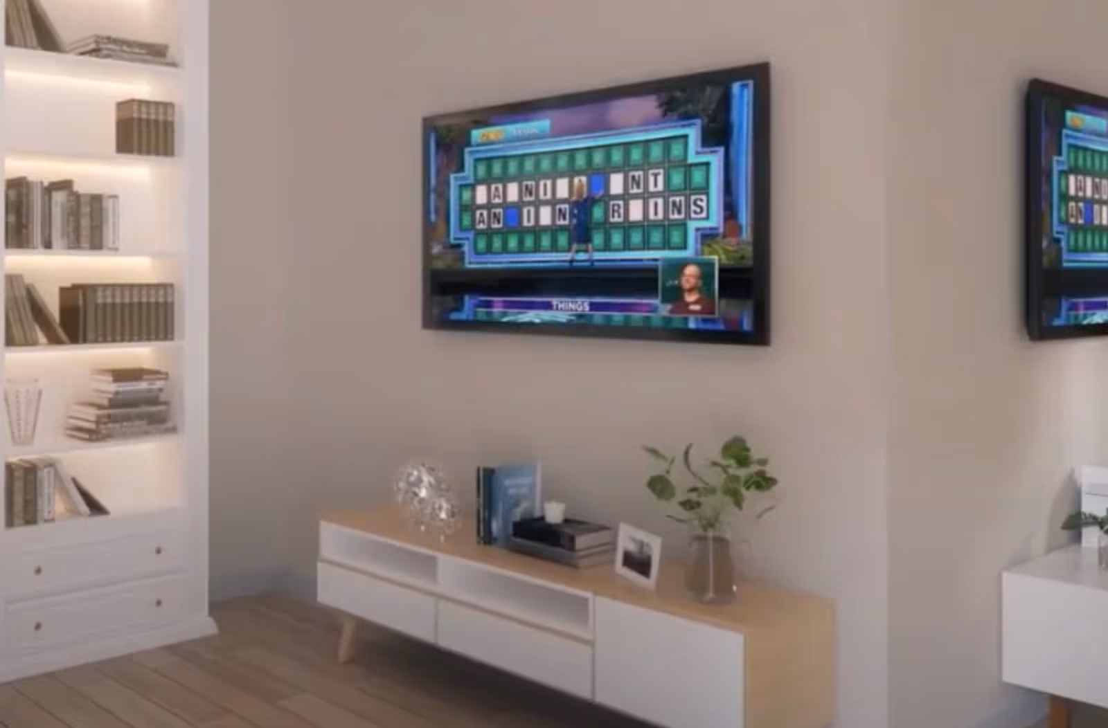 A modern living room area with a wall-mounted flat screen TV displaying a game show. Below the TV is a wooden and white media console with books, a decorative glass object, a small plant in a glass vase, and a framed photo. To the left, there is a built-in white bookshelf with several books and decorative items. The room has light-colored walls and wooden flooring.