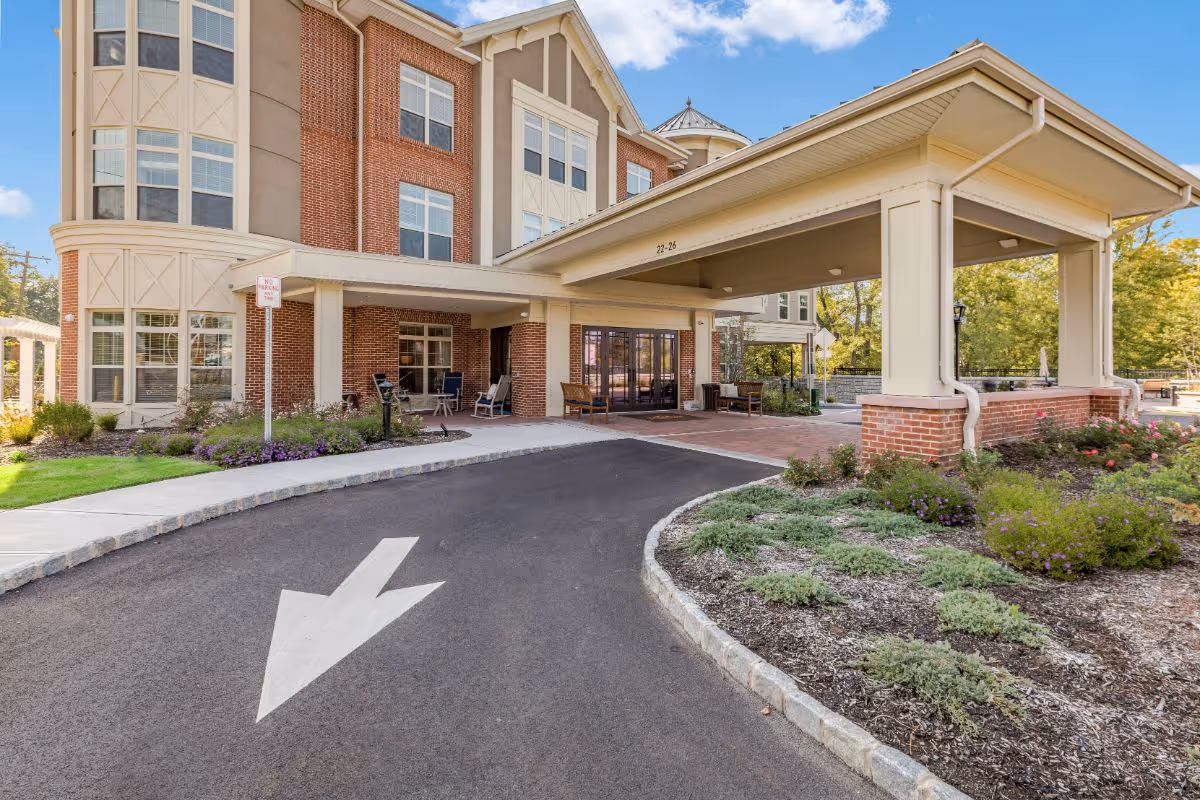 Covered porte-cochère drop-off and front entrance of a brick senior living building with a driveway arrow and landscaped beds.