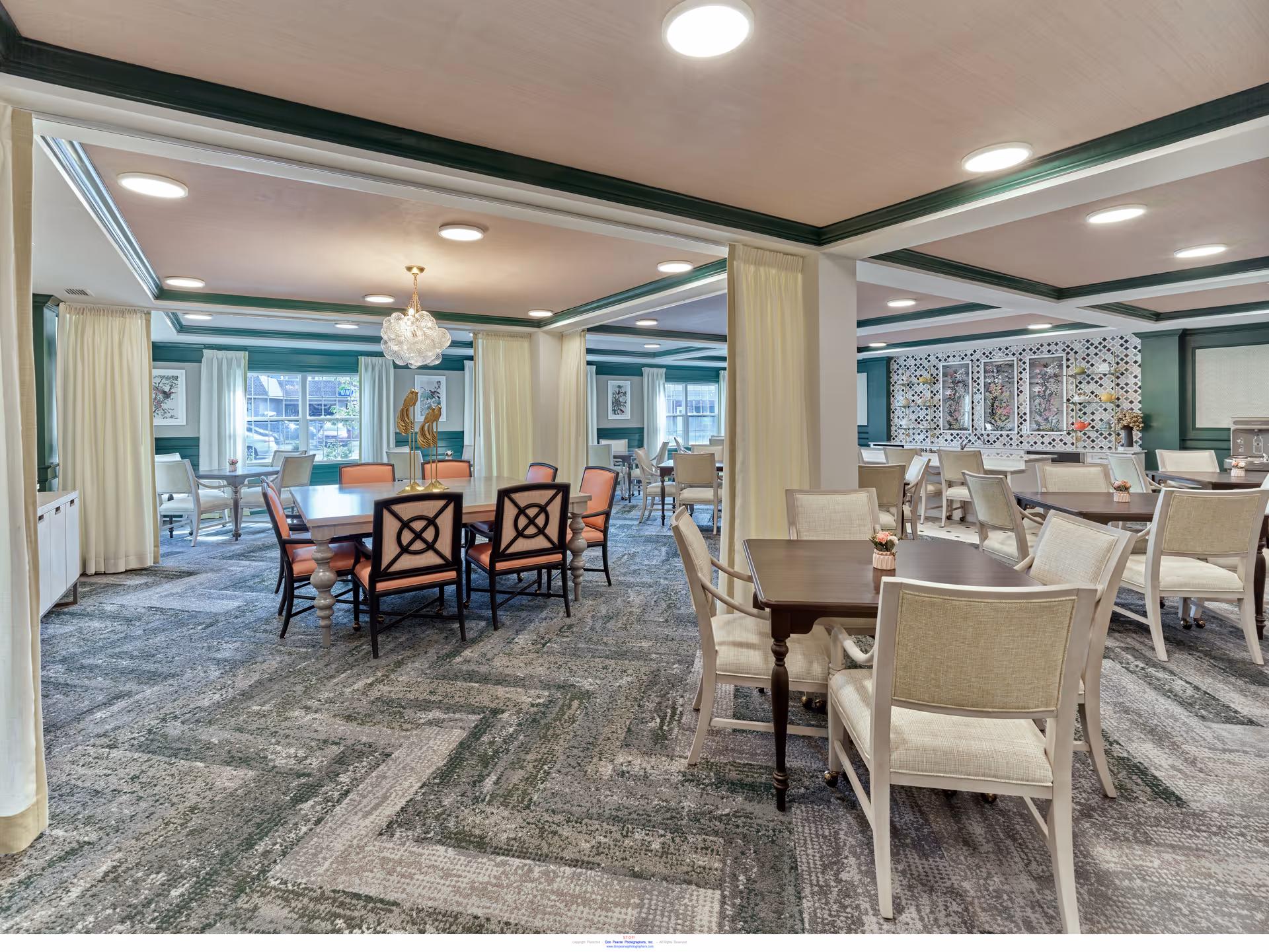 Spacious dining room with multiple tables and chairs arranged neatly. The room features a patterned carpet, green and white walls with large windows letting in natural light, and ceiling lights. There are decorative elements on the walls and a chandelier above one of the tables.
