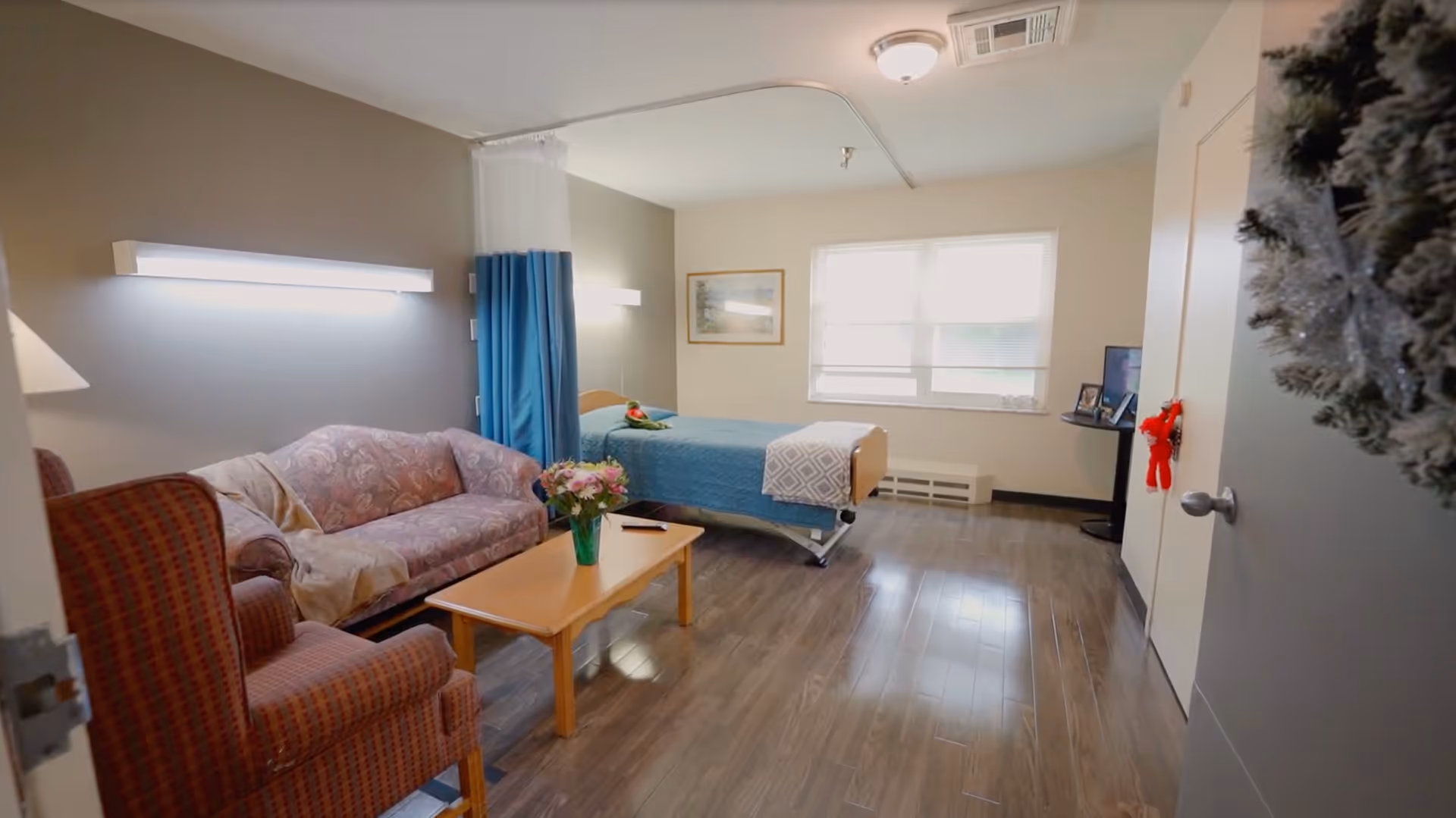 Private senior care room with a bed, sofa, armchair, coffee table with flowers, and a TV near a window.