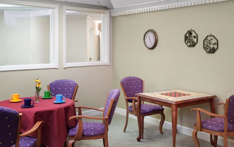 A cozy room with two tables and several purple cushioned chairs. One table is round with a burgundy tablecloth and colorful cups and saucers on top, along with a small vase of yellow flowers. The other table is square with a built-in checkerboard game. The walls are light green with a clock and decorative wall hangings. There are two large windows on one wall.