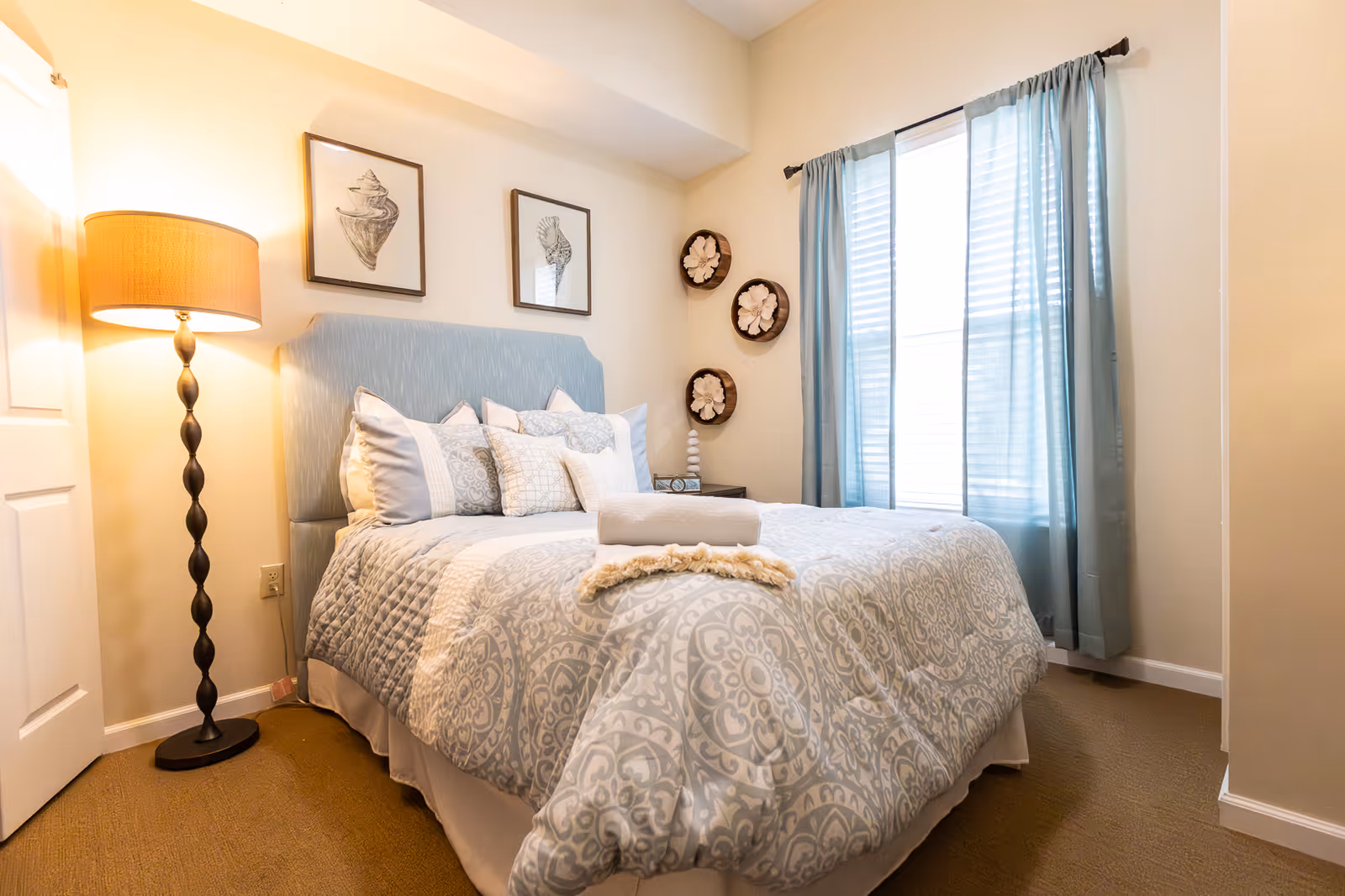 A cozy bedroom with a neatly made bed featuring patterned bedding and multiple pillows. There is a rolled towel and a decorative throw on the bed. To the left of the bed is a tall floor lamp with a warm light. Above the bed are two framed shell illustrations, and on the adjacent wall are three circular wall decorations. A window with light blue curtains allows natural light into the room.