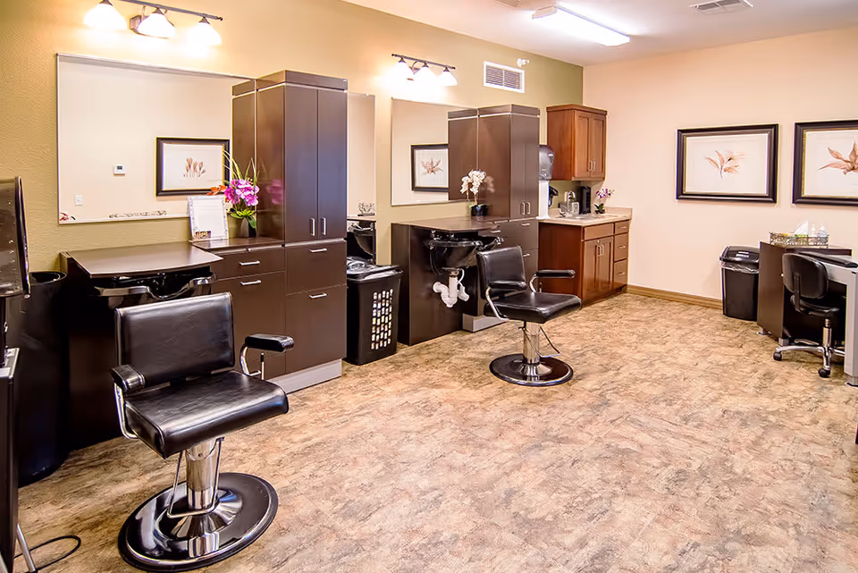 Interior view of a salon area in a senior living facility with two black salon chairs in front of sinks and large mirrors. The room has beige walls, wood cabinets, framed botanical artwork, and a desk with a chair in the corner.