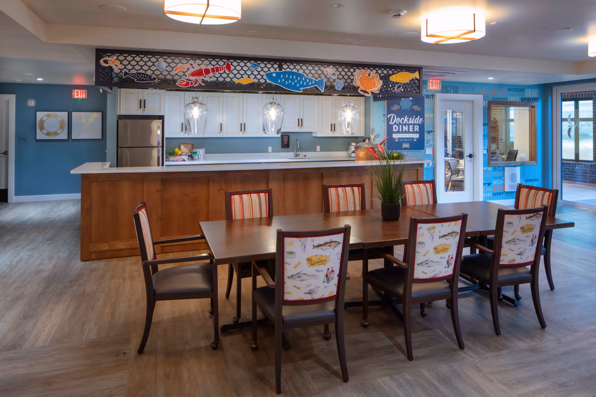 A dining area in Aegis Living Lake Union featuring a long wooden table with eight chairs, each with a fish-themed upholstery on the backrest. Behind the table is a kitchen counter with a wooden base and white countertop, illuminated by three hanging pendant lights. The back wall has white cabinets and a refrigerator, decorated with colorful fish and sea creature cutouts above. To the right, there is a blue wall with a sign that reads 'Dockside Diner' and a door leading to another room.
