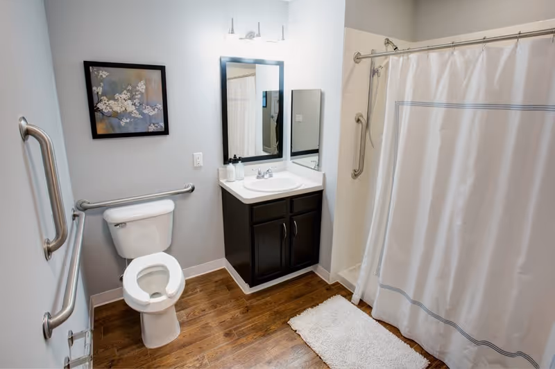 A clean bathroom with a white toilet, a dark wood vanity with a white sink, a mirror above the sink, a shower with a white curtain, and wooden flooring. There are grab bars near the toilet and shower for accessibility, a white bath mat on the floor, and a framed floral artwork on the wall.
