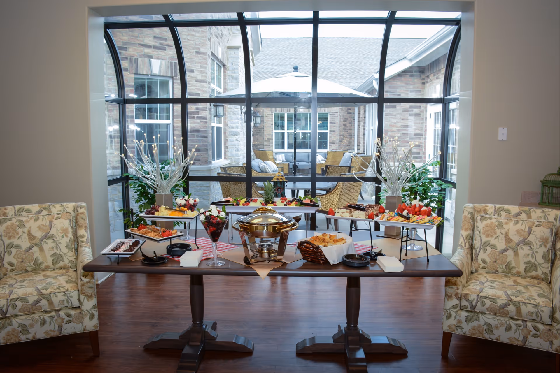 A buffet table set with various appetizers and desserts, including fruit platters, pastries, and a chafing dish, positioned between two floral upholstered armchairs. Behind the table is a large window with a view of an outdoor patio area with wicker furniture and an umbrella.