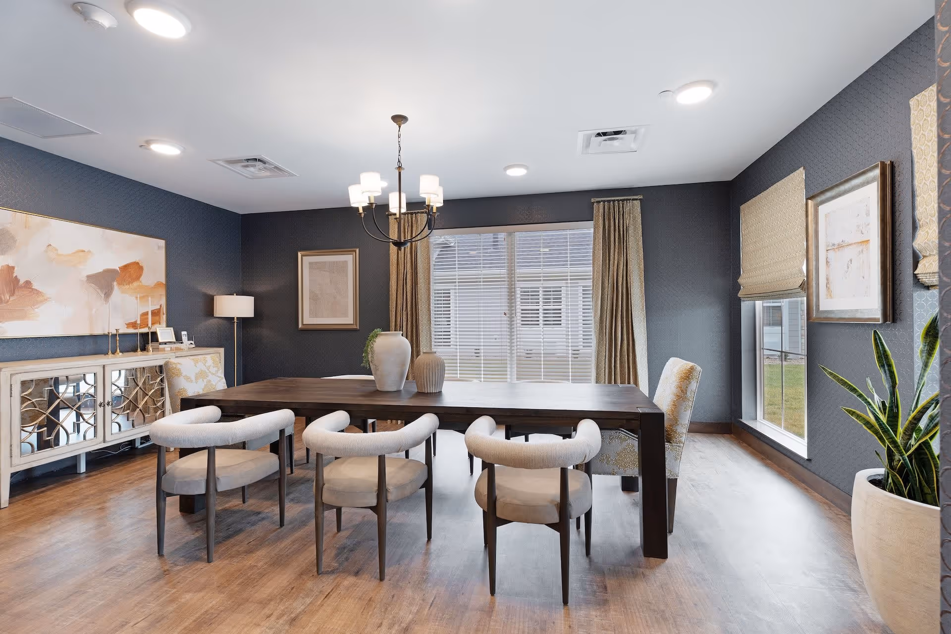 A modern dining room with a dark wooden table surrounded by six upholstered chairs, three on each side. The room has dark textured walls, large windows with beige curtains and blinds, a chandelier hanging above the table, and decorative vases on the table. There is a sideboard with mirrored doors and artwork on the walls, along with a potted plant near the window.