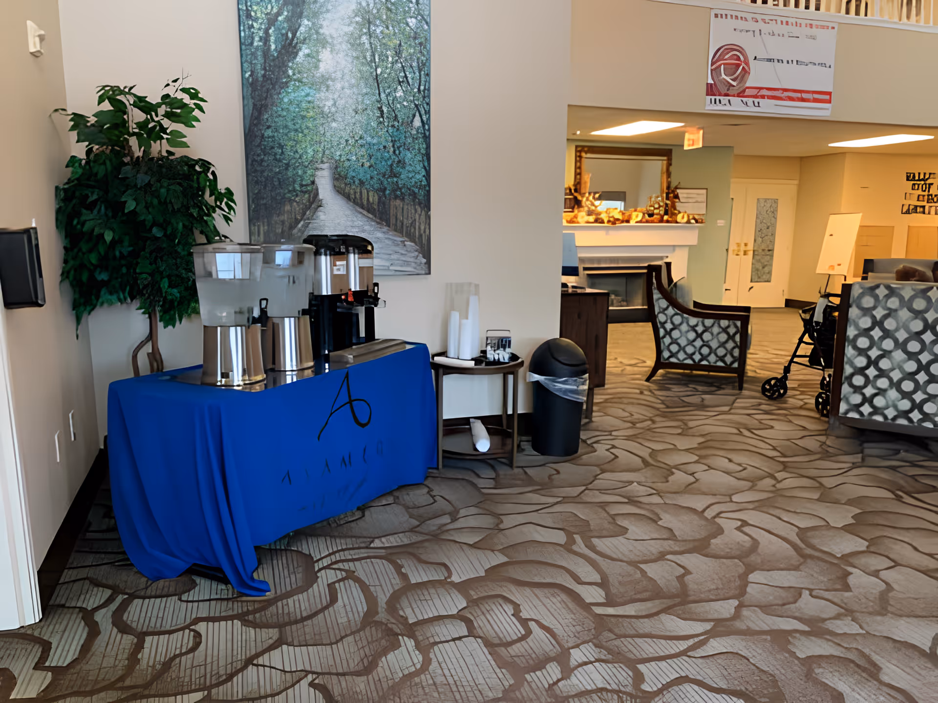 Interior view of a senior living facility lounge area with a beverage station covered by a blue tablecloth on the left, featuring water and coffee dispensers. There is a large painting of a forest pathway on the wall behind the beverage station. To the right, there are patterned armchairs, a fireplace with decorations on the mantel, and a walker near the seating area. The floor is carpeted with a textured pattern.