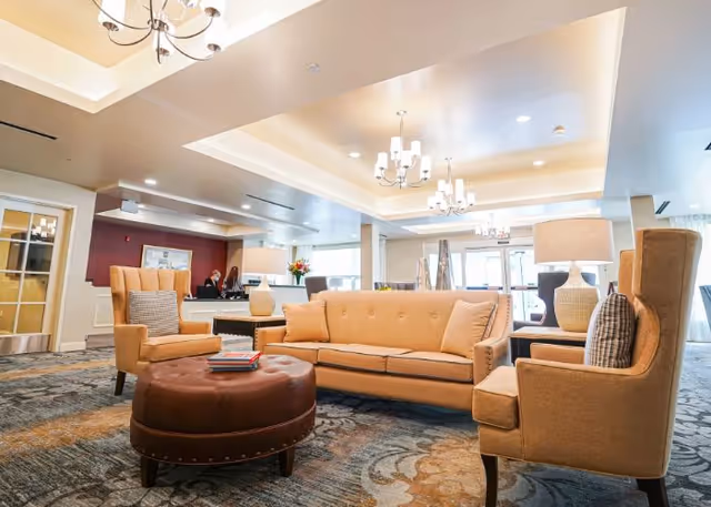 Bright, furnished lobby lounge with a beige sofa, matching armchairs, a round ottoman, and chandeliers under a recessed ceiling.