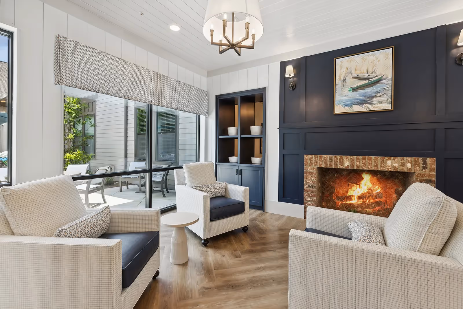A cozy living room area with three cushioned armchairs arranged around a small round side table. The room features a lit brick fireplace set in a dark blue paneled wall with a painting of a boat above it. Large windows with patterned valances provide a view of an outdoor patio area with chairs and greenery. The floor is wood with a herringbone pattern, and a modern chandelier hangs from the white paneled ceiling.